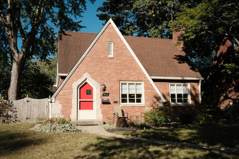 A red-brick cottage with a red front door, a lawn and mature shade trees